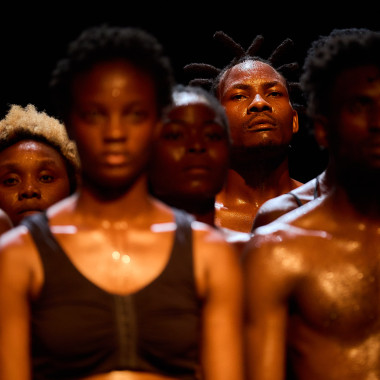 A scene from VAGABUNDUS: It shows several rows of dancers standing upright next to each other. The focus is on the person in the background looking into the camera.
