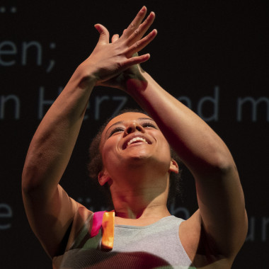 Ein Foto von Workshopleitung Naomi Sanfo-Ansorge. Es zeigt Naomi vor einer schwarzen Wand, auf der ein Text steht. Naomi trägt ein graues ärmelloses Tanktop und hält die Arme in die Luft. Dabei berühren sich ihre Hände.