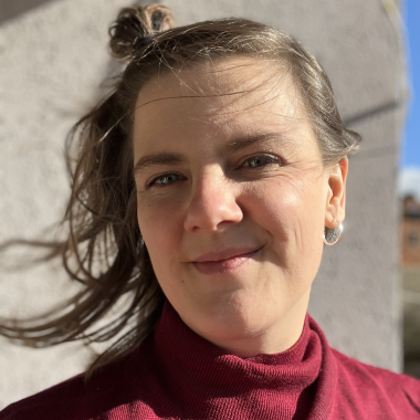 Ein Portrait von Workshopleitung Dr. Anna Wieczorek. Anna trägt einen roten Rollkragenpullover und hat braune länge Haare. Sie lächelt in die Kamera.