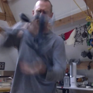 Ein Videostill der Arbeit TRI TEMPI (2023) von William Forsythe. Es ist ein Tryptichon und zeigt Forsythe stehend mit einem Objekt in der Hand.
