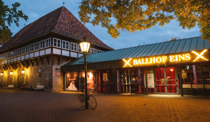 Das Ballhof Eins-Gebäude mit einem Schild mit der Aufschrift Ballhof Eins. Die gelbe Schrift ist hell erleuchtet. Das Schild befindet sich über den Eingang, der verglast ist. Vor dem Gebäude steht eine Laterne, die auch leuchtet.