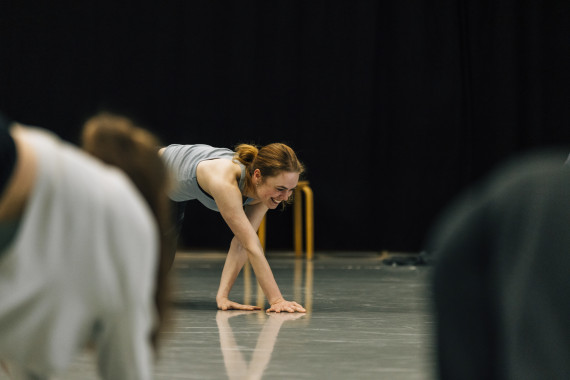 Ein Foto aus einem Workshop von Cara Rother. Es zeigt Cara Rother, die mit beiden Händen den Boden berührt und dabei lächelt. Im Bildvordergrund Workshopteilnehmer*innen von hinten. 