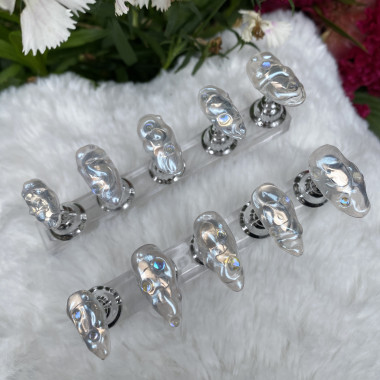 Zwei Reihen von Press-Ons von Nail Artistin Hong Ngoc Le, die auf einem weißen Plüschstoff liegen. Sie sind ovalförmig und silber. Auf den einzelnen Nägeln  sind organische Formen in der Farbe Silber zu sehen.