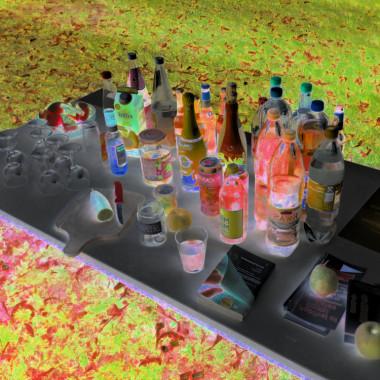 Negatively coloured image of an outdoor table with many bottles, glasses and drinks, surrounded by grassy ground depicted in bold, unnatural-looking shades of green and red.