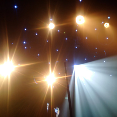 Several bright spotlights and small blue points of light shine against a dark background. Beams of light fan out into the room, creating an atmospheric stage or club atmosphere.