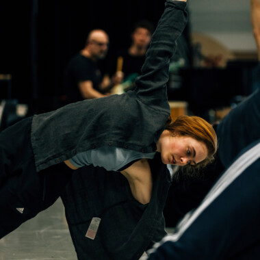 Eine Szene aus einem Workshop von Cara Rother. Es zeigt Cara Rother bei einer Art Dehnübung. Cara ist sportlich gekleidet und streckt die linke Hand in die Luft. 